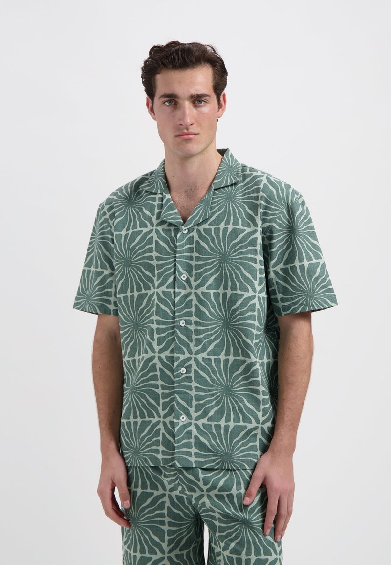 Korte mouwen shirt van groene stof met een getextureerd, bloemmotief. Inclusief een camp-col en knoopsluiting. Bijpassende broek zichtbaar.