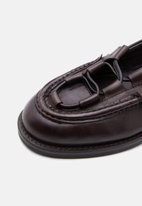 Σκούρο καφέ δερμάτινο μοκασίνι με στρογγυλεμένη μύτη, λεπτομερή ραφές και διακοσμητικά δεμένα λουριά στο επάνω μέρος του παπουτσιού.