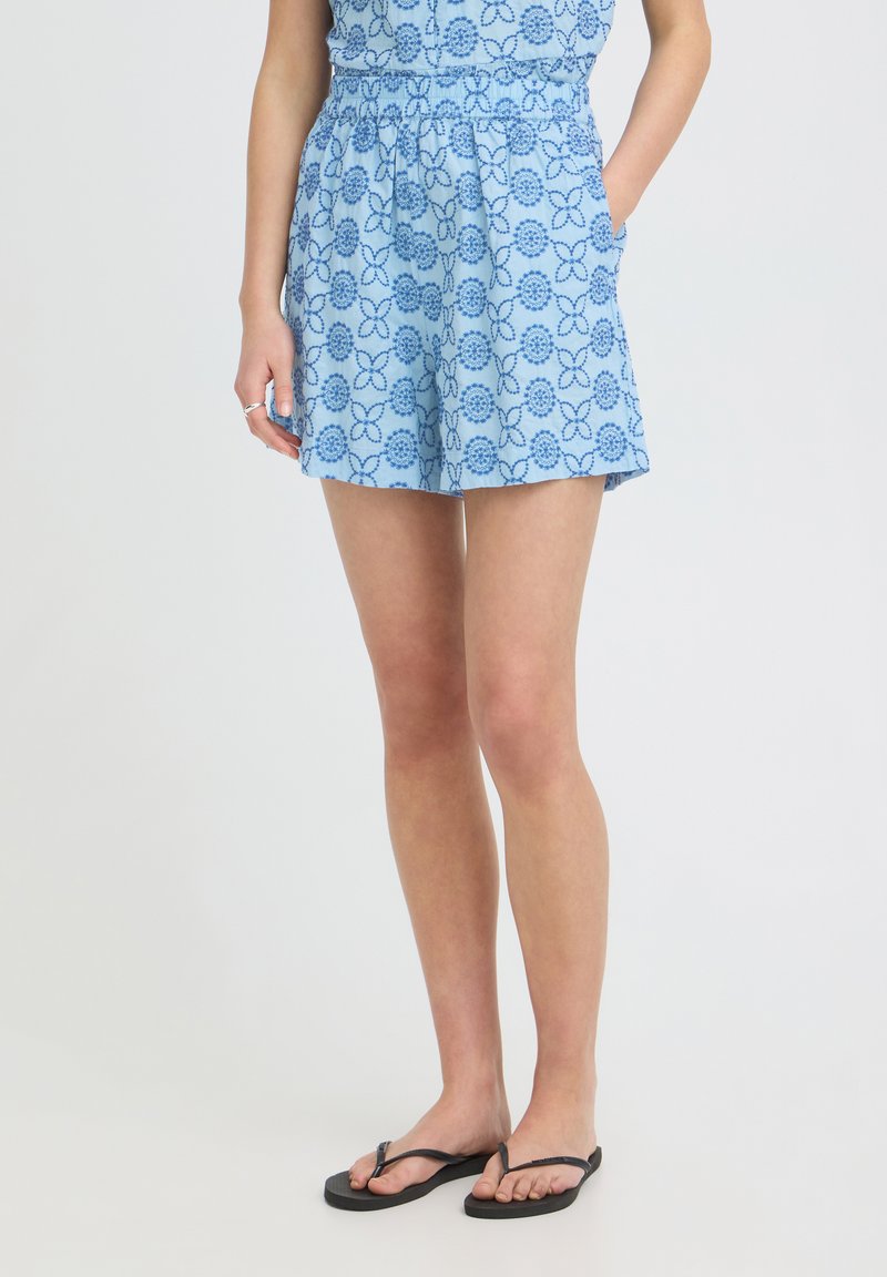 Femme portant un short bleu à motif floral avec une taille élastique, des poches et des tongs noires, debout devant un fond uni.