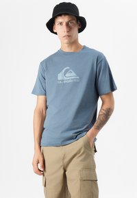 Ung mannlig modell iført en blå Quiksilver t-skjorte og beige cargo-shorts, stående mot en hvit bakgrunn, med en svart bøttehatt.