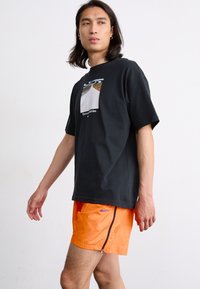 Svart t-skjorte med korte ermer og grafikk av en vei på forsiden samt tekst, paret med oransje nylonshorts med glidelås på siden og Nike-logo.