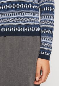 Gros plan d'une personne portant un pull à motifs bleu, blanc et noir avec un pantalon gris, montrant sa main droite sur le côté.
