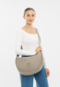 Szürke, kerek formájú üzenet táska, textil anyagból, állítható pánttal és logóval, egy fehér hosszúujjú pólót viselő személy által tartva.