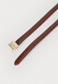 Barna bőr öv, több állítható lyukkal és arany tónusú négyzetes csattal, amelyen ívelt betűtípussal a logó van gravírozva, fehér háttér előtt.