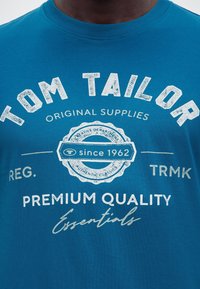 Μπλε πουκάμισο με λευκό κείμενο «Tom Tailor», «Original Supplies», «Since 1962», «Premium Quality Essentials» και γραφικό έμβλημα στο στήθος.