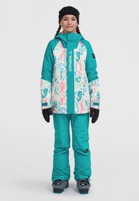 Turkis skijakke med et farverigt, abstrakt print og en frontlynlås, parret med matchende bukser. Sorte handsker og en hue fuldender outfittet.