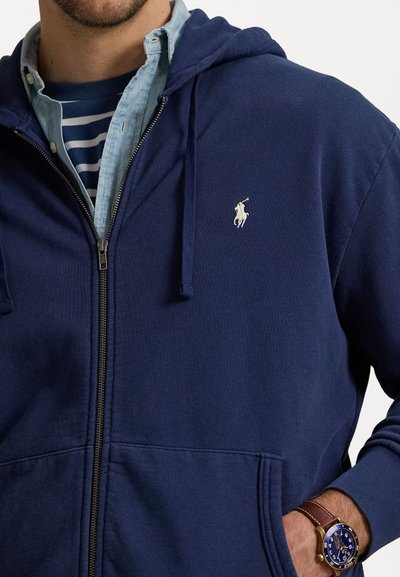Marinblå zip-up hoodie med dragsko, som har en vit broderad logga på vänster bröst och ett mjukt, texturerat tyg.