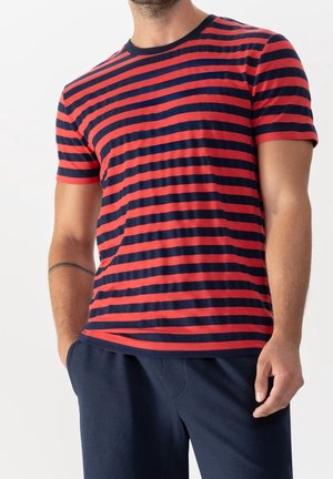 Man met een rood- en donkerblauw horizontaal gestreept shirt met korte mouwen en donkerblauwe broek, met één hand in de zak en een tatoeage op zijn linkerarm.
