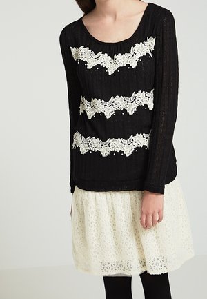 Frau trägt ein schwarzes Langarmshirt mit drei horizontalen weißen Spitzenstreifen mit Blumenmuster und einen weißen Spitzenrock.