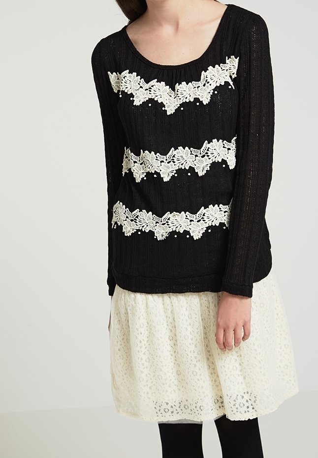 Femme portant un haut noir à manches longues avec trois bandes horizontales de dentelle blanche à motifs floraux et une jupe en dentelle blanche.