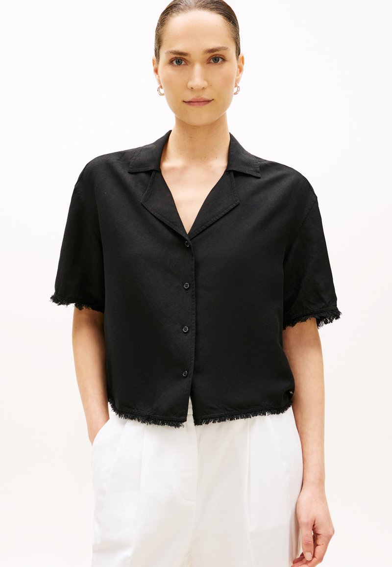 Mujer con una camisa negra de manga corta y botones, con bordes desgastados, y pantalones blancos de talle alto, de pie contra un fondo liso.