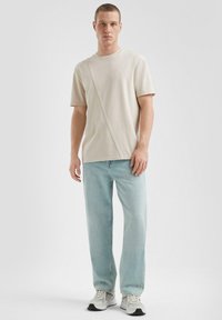 Jasno beżowa koszulka z ukośnym szwem, zestawiona z jasnoniebieskimi dżinsami. Buty są białe z szarymi akcentami. Prosty, minimalistyczny design.