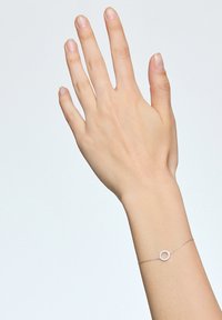 Srebrna bransoletka z okrągłym zawieszką ozdobioną małymi przezroczystymi kamieniami, wyeksponowana na dłoni na jasnym tle.