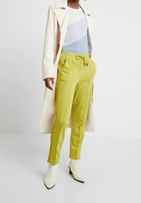 Lime green tapered pants with a drawstring waist, paired with a light blue and white color-blocked top and a cream coat. White heeled boots.