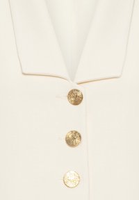 Gros plan d'un manteau couleur crème avec trois grands boutons dorés texturés et un large col à revers.