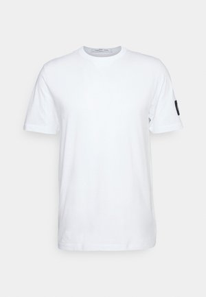 T-shirt blanc à manches courtes avec un petit patch noir sur la manche gauche, présenté sur un fond clair uni.