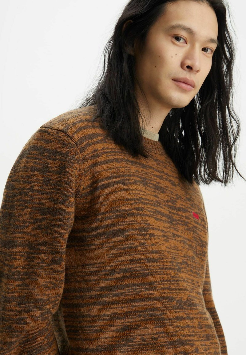 Suéter de punto marrón con patrón, cuello redondo y mangas largas. Presenta un sutil logo rojo en el pecho y un acabado texturizado.