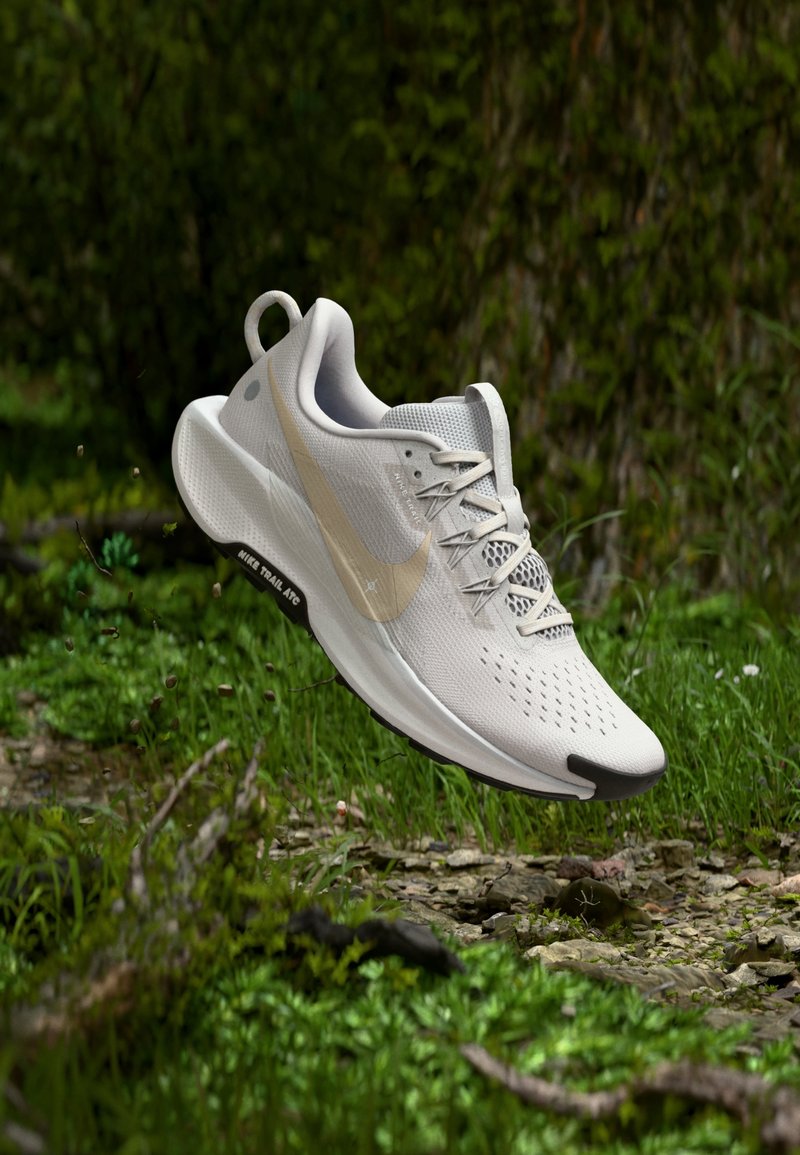 Chaussure de course Nike blanche et beige suspendue au-dessus d'un sol forestier rocheux avec des plantes vertes et des arbres flous en arrière-plan.