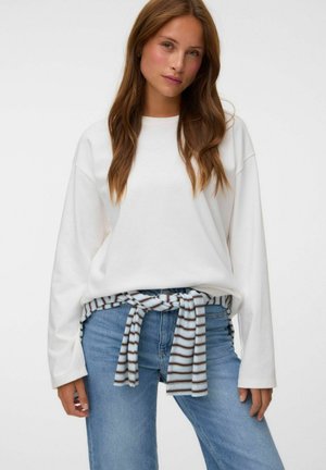 Jeune femme aux longs cheveux bruns porte une chemise blanche ample à manches longues, un jean bleu clair et une chemise rayée nouée autour de la taille.