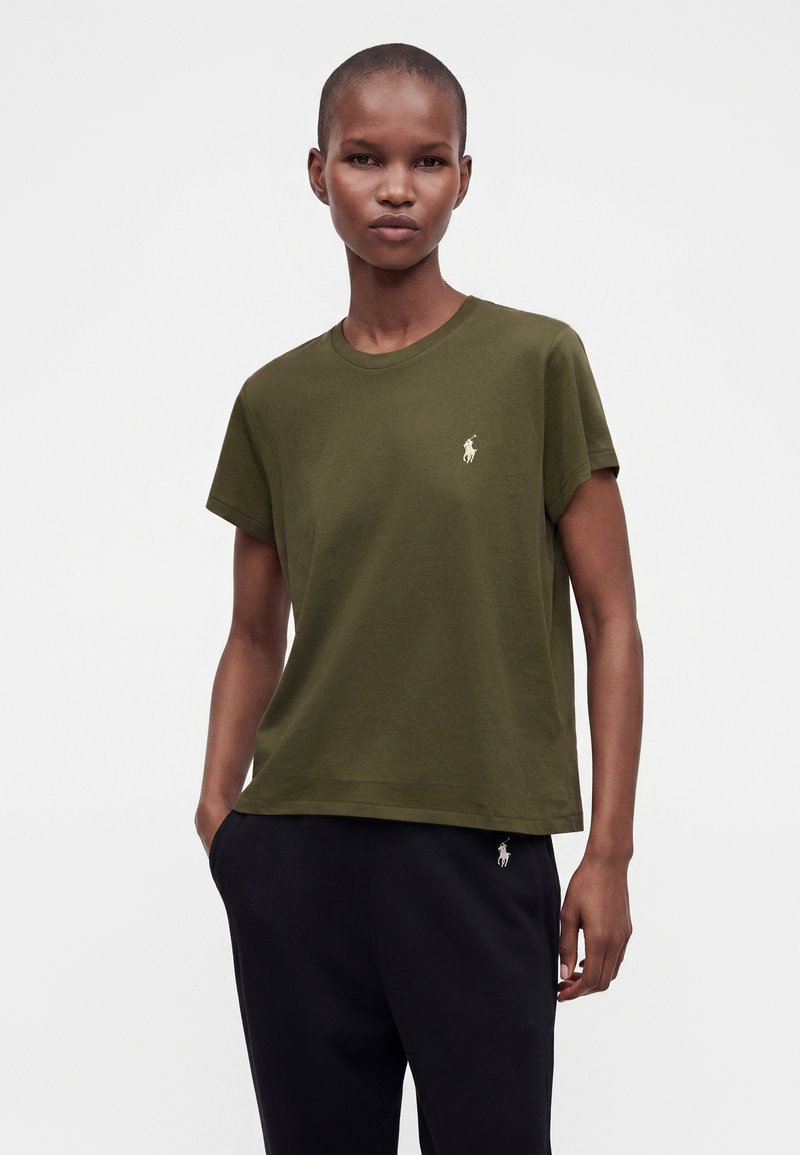 Personne mince portant un t-shirt vert olive à manches courtes et un pantalon noir, les mains dans les poches, debout devant un fond blanc uni.