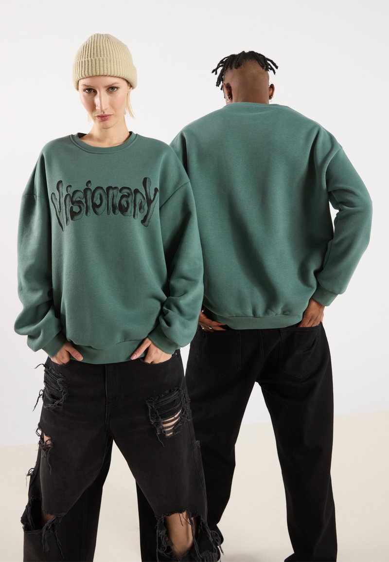 Felpa verde con la scritta "Visionary" in nero corsivo. Vestibilità rilassata, polsini a coste, indossata con jeans neri strappati. Modello con un berretto beige.