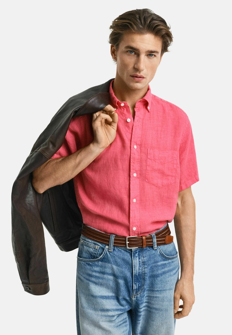 Kurzärmliges pinkes Leinenhemd mit Button-Down-Kragen, kombiniert mit blauen Jeans und einem braunen Ledergürtel, während eine dunkle Jacke gehalten wird.