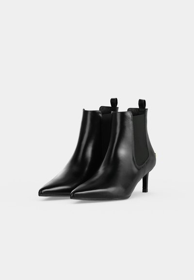 Une paire de bottines en cuir noir à bouts pointus, avec des empiècements latéraux élastiques, des tirettes et des talons fins, sur un fond uni.