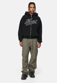 Felpa nera con zip e logo bianco, abbinata a pantaloni cargo larghi di colore verde oliva. Il modello indossa sneakers nere, dotate di molteplici tasche cargo.