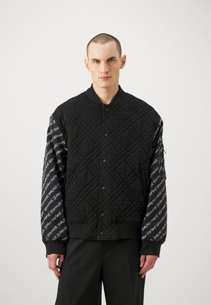 Gepolsterte schwarze Bomberjacke mit solidem Vorderteil und gemusterten Ärmeln, die weißen Text tragen. Rippbündchen und Kragen runden das Design ab.