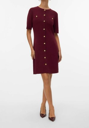 Femme portant une robe bordeaux mi-longue à manches courtes avec des boutons dorés sur le devant et sur les poches poitrine, associée à des talons bordeaux.