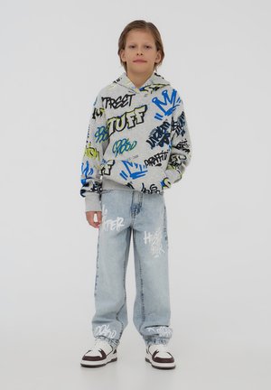 Felpa grigia con testo in stile graffiti multicolore, abbinata a jeans in denim blu chiaro con design di testo bianco. Sneakers bianche completano il look.