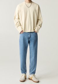 Personne portant une chemise crème à manches longues, un jean bleu clair, des baskets blanches avec des rayures orange, et un collier en chaîne argentée, les mains dans les poches.