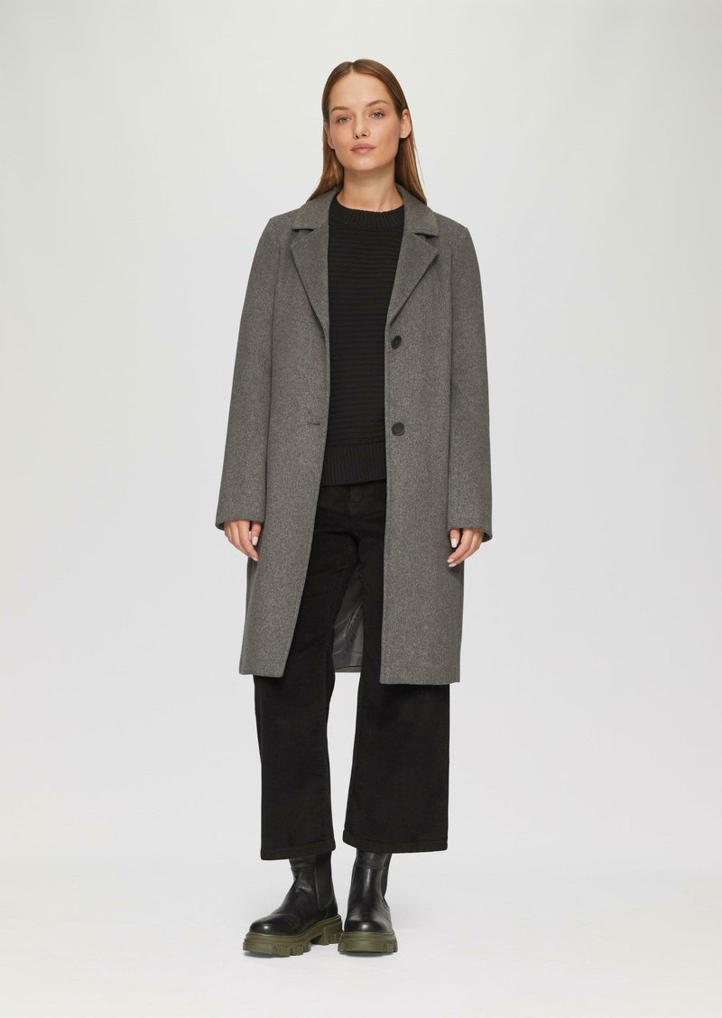 Manteau en laine grise avec un col à revers, fermeture à bouton et coupe droite. Porté sur un pull noir à côtes et un pantalon noir à large jambe. Bottes noires.