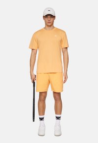 Orange atletisk t-shirt och shorts, bomullsblandat tyg, matchat med vit keps och vita sneakers. Hållande en svart tennisracket.