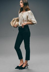 Blusa con bordado floral en crema y oro, jeans ajustados negros y tacones negros tipo slingback con un lazo. Sosteniendo un clutch metálico dorado.