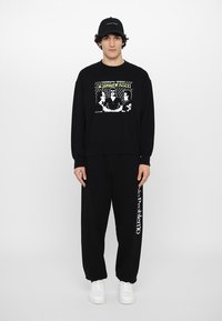 Black sweatshirt and pants set, featuring a graphic print on the sweatshirt. White text on the pant leg. Finished with white sneakers and a cap.