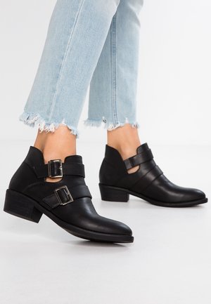 Sorte læderankelstøvler med dobbelt spænde, båret med lyseblå jeans med frynset kant, vist på hvid baggrund.