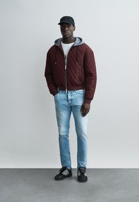 Noor mees, kes kannab burgundia värvi tikitud jopet, helesiniseid teksapükse, musta nokamütsi ja musti tenniseid, seisab tavalise halli tausta ees.