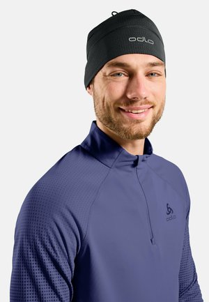 Uomo che indossa un berretto Odlo nero e una maglia Odlo blu a manica lunga con zip a quarto, sorridendo su uno sfondo chiaro e semplice.
