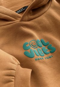 Brun hættebluse med blød struktur, der har et teal tryk med teksten "COOL VIBES" og et smilende ansigt. Inkluderer ribbede manchetter og hætte.