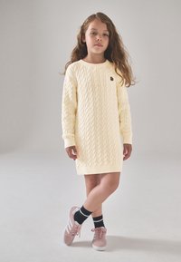 Mezgimo su kabelio raštu suknelė švelnios geltonos spalvos su ilgomis rankovėmis, papuošta mažyte emblema. Derinama su rožiniais batais ir juodomis kojinėmis.