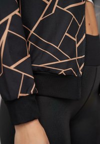 Schwarze Sportjacke mit einem geometrischen, roségoldenen Muster, Reißverschluss vorne und gerippten Bündchen, kombiniert mit einfarbigen schwarzen Leggings.