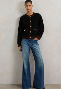 Černý pletený cardigan s hnědými akcenty a zlatými knoflíky, kombinovaný s rozšířenými modrými džíny z denimu s předními švy a vybledlým vzhledem.