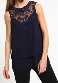 Blouse sans manches bleu marine avec un panneau en dentelle transparente orné de broderies florales au niveau de l'encolure, ourlet à volants et finition en tissu lisse.