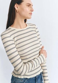 Beżowy dopasowany top z długim rękawem w poziome granatowe paski i falbaniastym dołem, w komplecie z niebieskimi dżinsami. Widoczne złote kolczyki w kształcie kół.