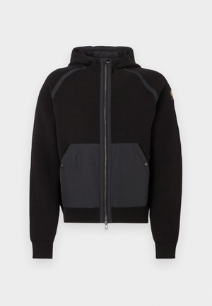 Felpa nera con cappuccio, caratterizzata da un pannello frontale opaco nero a contrasto, chiusura con zip e tasche laterali con bottoni a pressione.