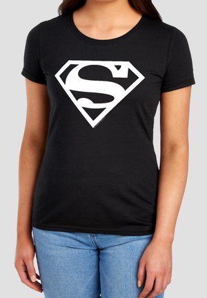 Woman wearing a black fitted t-shirt with a large white Superman logo and light blue jeans standing against a plain background.