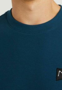 Mörk teal sweatshirt med textur, rund hals och en liten svart etikett med en vit logotyp. Tyget har ett vävt mönster.