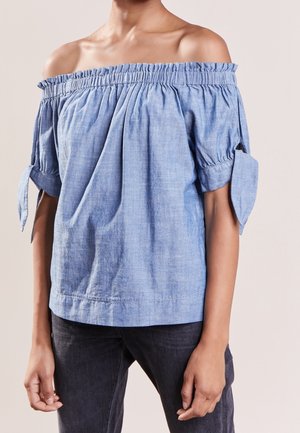 Vrouw draagt een lichtblauwe off-shoulder top met korte mouwen, gestrikt in strikjes, en een donkere broek tegen een effen achtergrond.