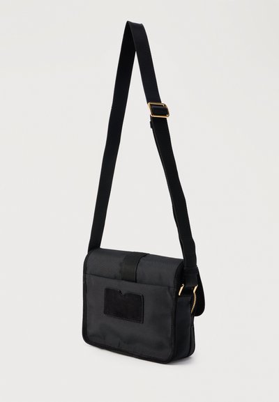 Μαύρη ορθογώνια τσάντα χιαστί με ρυθμιζόμενο ιμάντα, μπροστινή θήκη για κάρτες και εξαρτήματα σε χρυσό τόνο σε ανοιχτό φόντο.